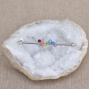 May include: A rainbow colored surgical steel barbell with a pointed end. The barbell is adorned with seven small round opal gems in a rainbow color scheme.