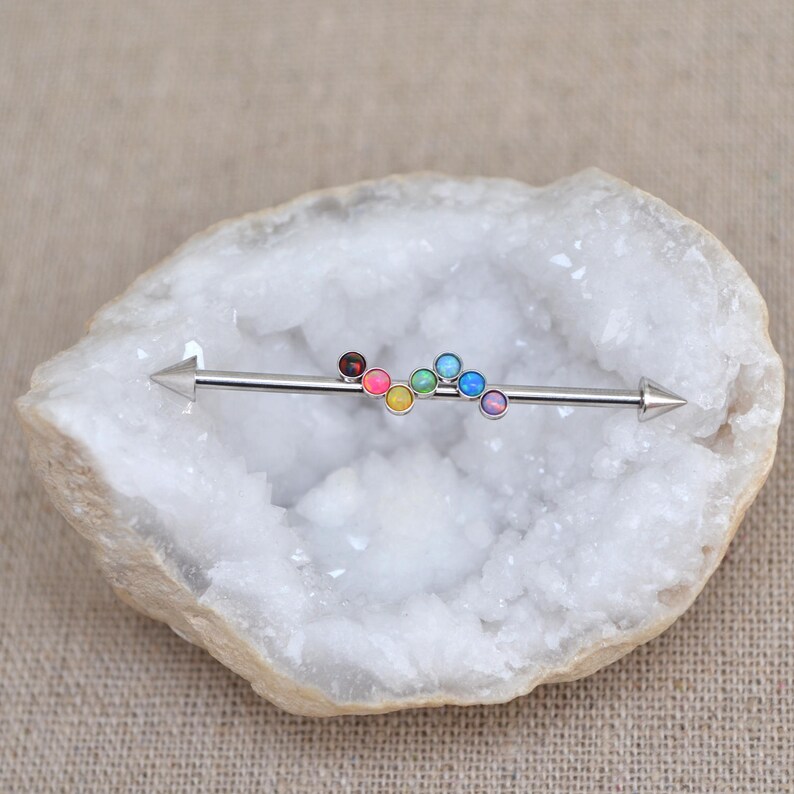 May include: A rainbow colored surgical steel barbell with a pointed end. The barbell is adorned with seven small round opal gems in a rainbow color scheme.
