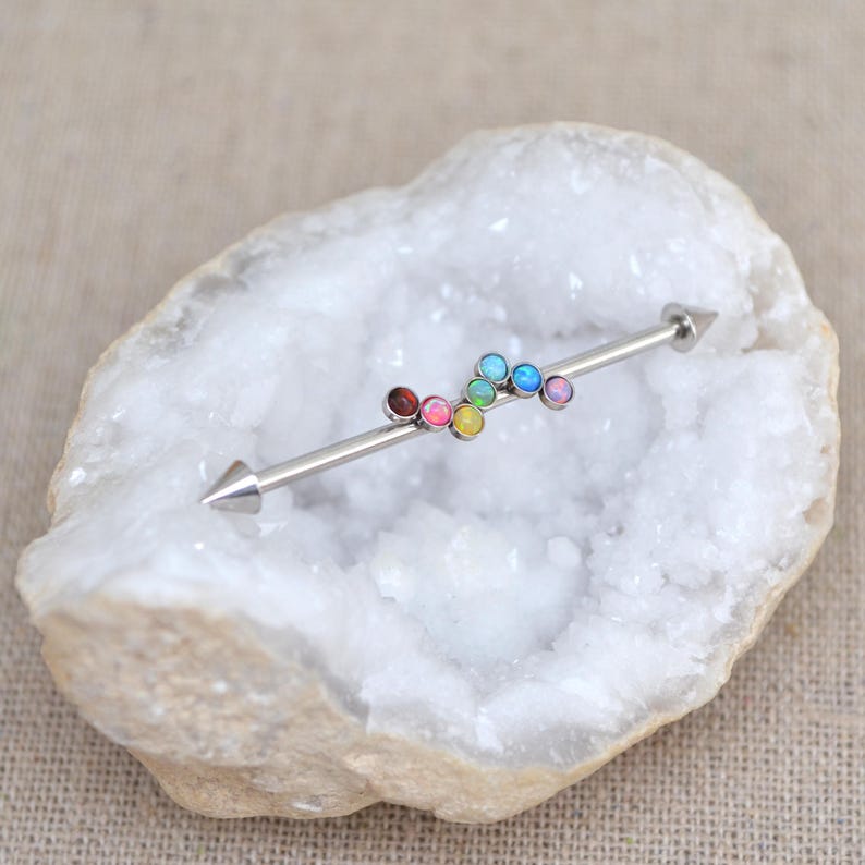 May include: A rainbow colored opal barbell with a silver bar and pointed ends. The barbell is set on a white crystal background.