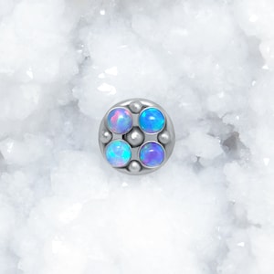 May include: A circular silver-toned body jewelry piece featuring four round, iridescent blue and purple cabochon stones arranged in a square pattern, accented by small metal spheres between the stones and at the center of the setting.