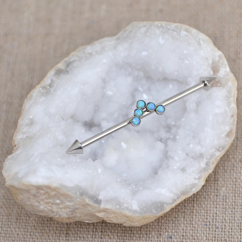 Puede incluir: Un piercing industrial de barra plateada con dos extremos puntiagudos y un grupo central de cuatro peque&ntilde;as piedras redondas de color azul claro, similares a &oacute;palos, dispuestas en un patr&oacute;n triangular sobre la barra.
