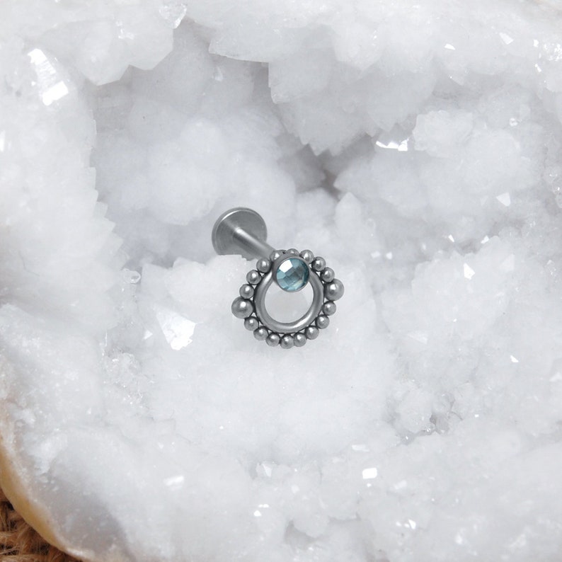 May include: Silver body jewelry with a small blue gemstone and a beaded circle design.