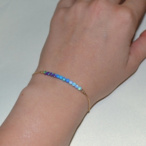 Puede incluir: Un delicado brazalete de cadena dorada con una fila de pequeñas cuentas de ópalo iridiscente en tonos de morado, azul y blanco.