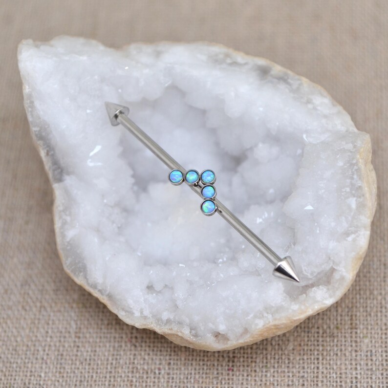 Puede incluir: Una pieza de joyer&iacute;a corporal tipo barra industrial en tono plateado que presenta una barra met&aacute;lica recta con extremos puntiagudos y un grupo de cuatro peque&ntilde;as piedras redondas de color azul claro iridiscente.