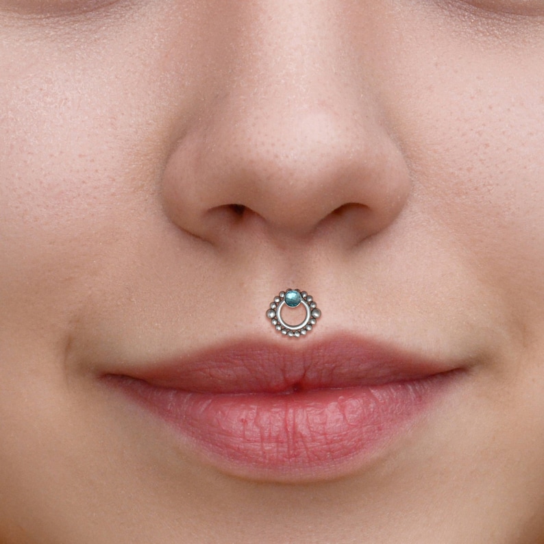 May include: A silver lip ring with a small turquoise gem in the center. The ring is made of small beads and has a circular design.