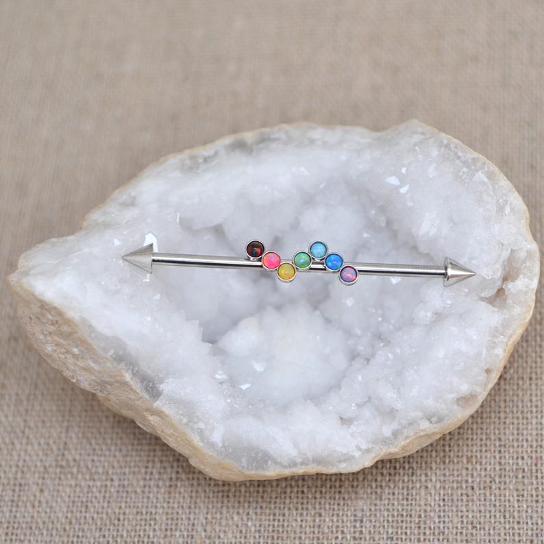 May include: A silver barbell with a rainbow of opal gemstones. The barbell is on a white crystal background.