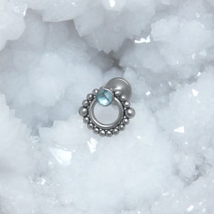 May include: A silver body jewelry piece with a circular design and a light blue gemstone. The jewelry is set against a white background.