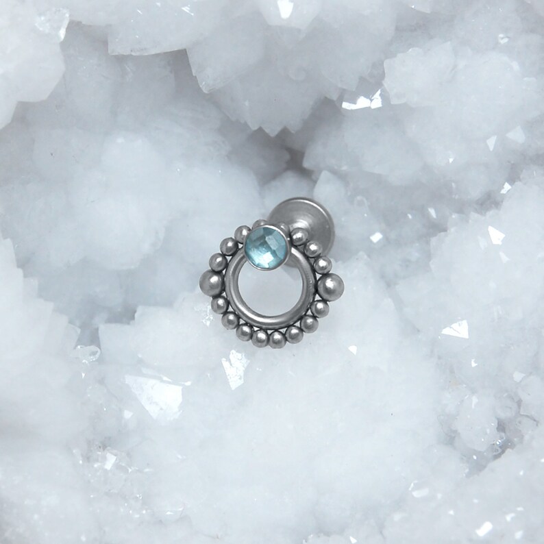 May include: A silver body jewelry piece with a circular design and a light blue gemstone. The jewelry is set against a white background.