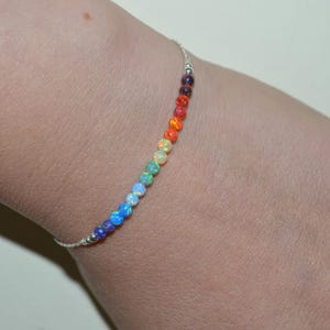 May include: A delicate bracelet featuring a single row of small, round, multi-colored opal beads arranged in a spectrum of colors including red, orange, yellow, green, blue, and purple, strung on a thin, metallic silver-toned chain.