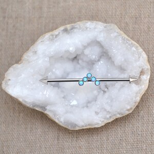 Puede incluir: Una joya corporal tipo barra industrial de tono plateado con un adorno central en forma de chevr&oacute;n compuesto por cinco peque&ntilde;as piedras redondas de color azul claro que recuerdan al &oacute;palo. La barra tiene extremos c&oacute;nicos en ambos lados.