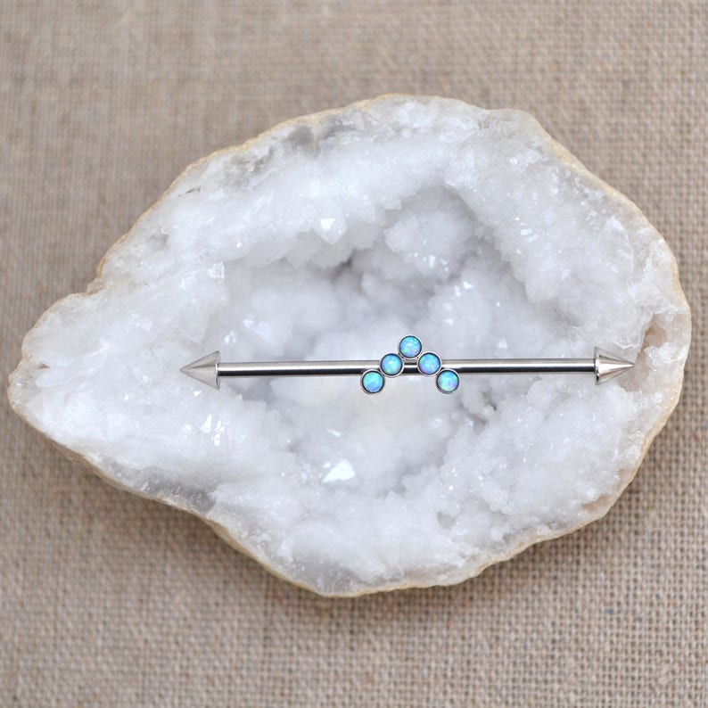 Puede incluir: Una joya corporal tipo barra industrial de tono plateado con un adorno central en forma de chevr&oacute;n compuesto por cinco peque&ntilde;as piedras redondas de color azul claro que recuerdan al &oacute;palo. La barra tiene extremos c&oacute;nicos en ambos lados.