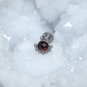 May include: A silver-toned metal body jewelry stud featuring a central round faceted dark red gemstone. The setting is accented with a series of small metal spheres arranged in a semi-circular pattern around the base of the stone, attached to a flat circular backing post.