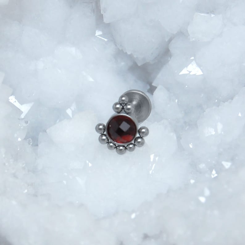 May include: A silver-toned metal body jewelry stud featuring a central round faceted dark red gemstone. The setting is accented with a series of small metal spheres arranged in a semi-circular pattern around the base of the stone, attached to a flat circular backing post.