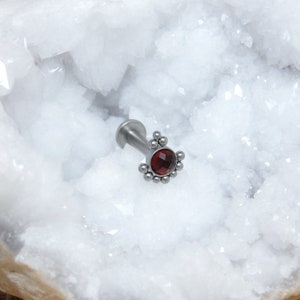 May include: A silver-toned labret stud body jewelry piece featuring a circular, faceted deep red gemstone at the center. The setting is adorned with a decorative arc of small metal spheres surrounding the stone, attached to a flat back post.
