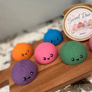 Puede incluir: Un conjunto de cinco cake pops coloridos con forma de bola de masa en morado, naranja, rosa, azul y verde, cada uno con una pequeña cara sonriente con ojos y boca negros. Se observa un recipiente redondo de cartón con la etiqueta The Sweet Duo.