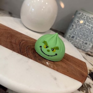 May include: A light green, dome-shaped chocolate treat featuring a hand-piped face with yellow eyes, dark green eyebrows, and a thin black smile. The confection has a pointed top, resembling a stylized character design with smooth icing.
