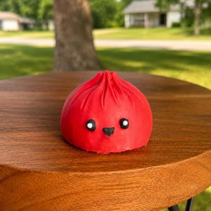 May include: A small, bright red, rounded dumpling-shaped figurine with a pleated top. It features a simple face consisting of two small white circular eyes with black pupils and a tiny black triangular mouth centered on the front surface.