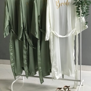 May include: Four satin robes hanging on a white rack. Three robes are sage green and one is white. The white robe has the word "Bride" embroidered on the back in gold thread.