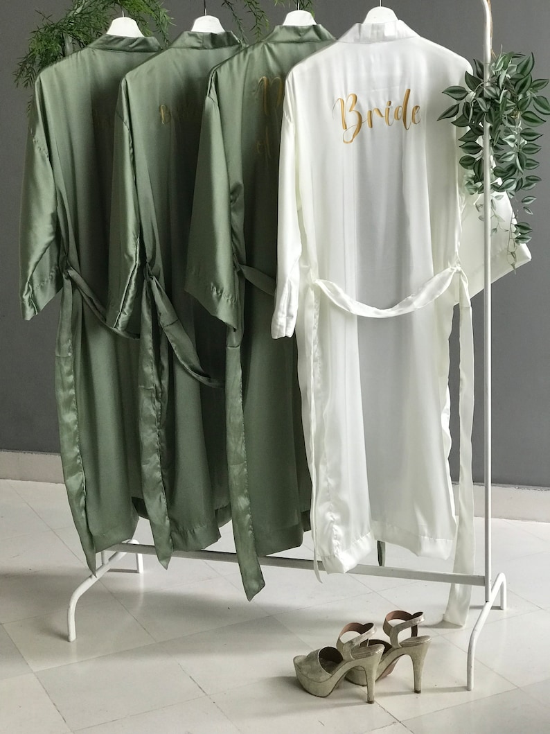 May include: Four satin robes hanging on a white rack. Three robes are sage green and one is white. The white robe has the word "Bride" embroidered on the back in gold thread.