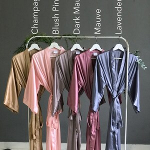 May include: Six satin robes in different colors hanging on a rack. The robes are in the colors champagne, blush pink, dark mauve, mauve, lavender gray.