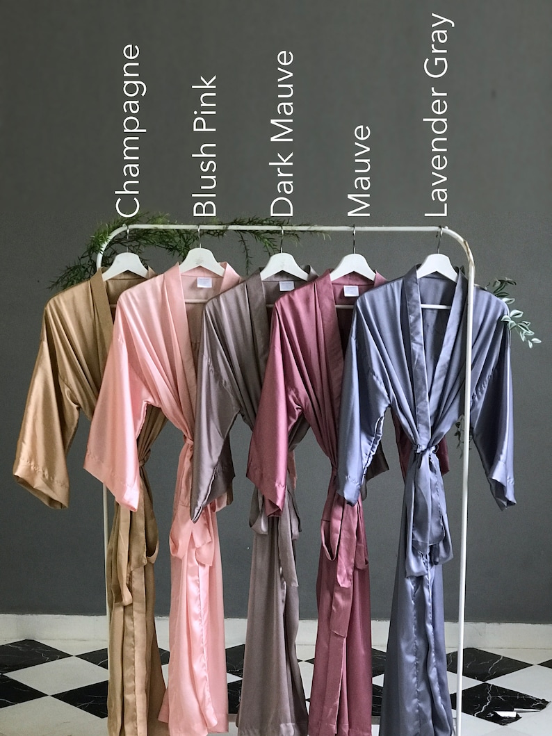 May include: Six satin robes in different colors hanging on a rack. The robes are in the colors champagne, blush pink, dark mauve, mauve, lavender gray.