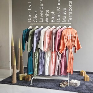 May include: A rack of 10 satin robes in various colors, including dark teal, olive, lavender gray, white, blush, dark mauve, mauve, and terracotta. The robes are hanging on a white metal rack with a white background.