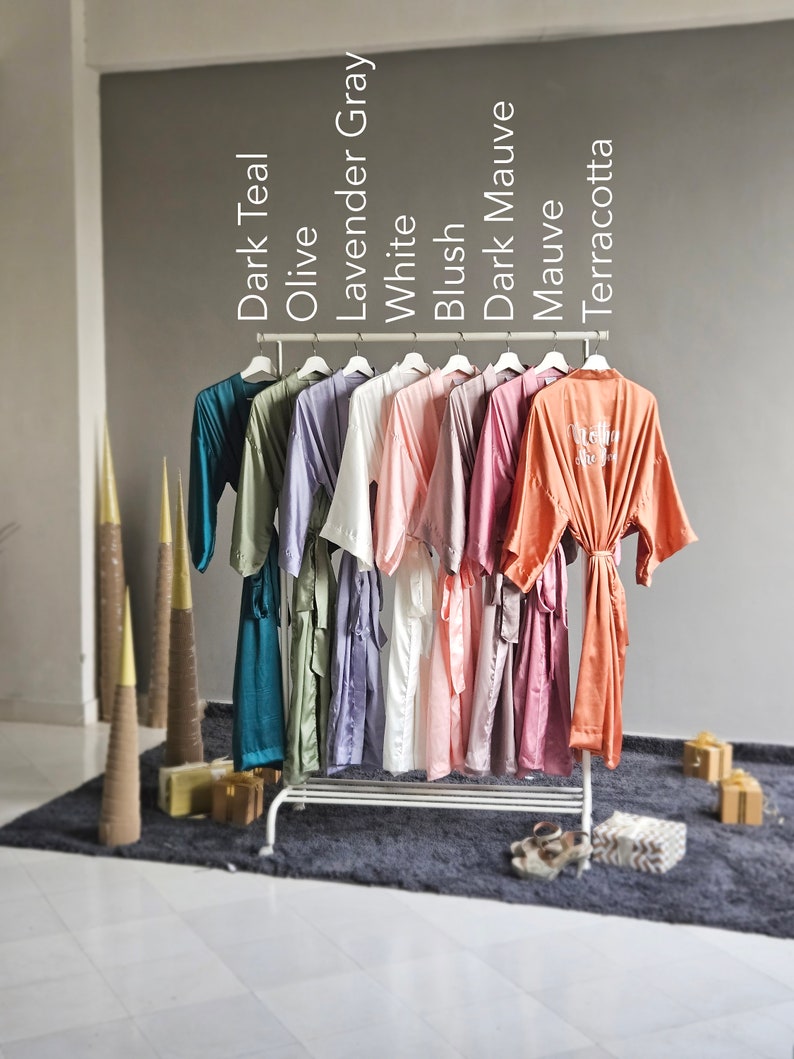 May include: A rack of 10 satin robes in various colors, including dark teal, olive, lavender gray, white, blush, dark mauve, mauve, and terracotta. The robes are hanging on a white metal rack with a white background.