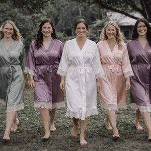 May include: Five women wearing matching satin robes in various colors: sage green, lavender, white, peach, and plum. Each robe has a lace trim and a matching belt. The robes are personalized with names embroidered in gold thread.