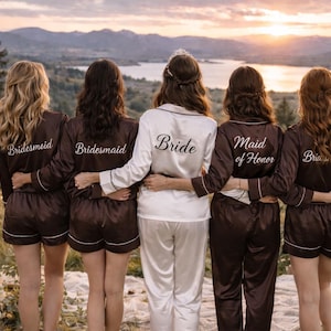 Könnte beinhalten: Fünf Frauen in passenden Pyjamas stehen draußen zusammen. Vier tragen braune Satin-Pyjamas mit weißer Paspelierung und dem Text "Bridesmaid" oder "Maid of Honor" auf dem Rücken. Die Braut trägt einen weißen Pyjama mit "Bride" auf dem Rücken.