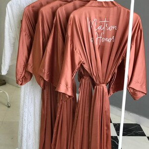 a row of brown satin robes hanging on a rack, with the backs of the robes featuring the text "Matron & Honor" printed on them.