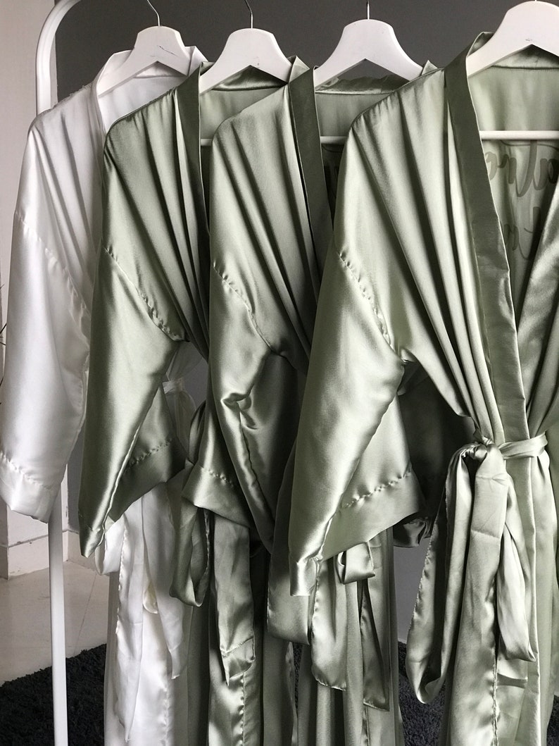 May include: Three satin robes, two in a light sage green color and one in white. The robes have long sleeves and a sash tie at the waist. The sage green robes have a subtle sheen and are displayed on hangers.
