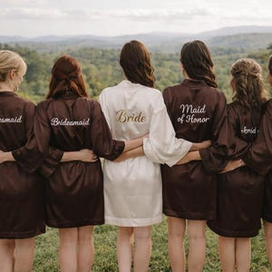 Könnte beinhalten: Eine Gruppe von Personen, die passende Roben tragen, steht im Freien mit dem Rücken zur Kamera. Die Roben sind braun, bis auf eine weiße Robe. Auf den Roben steht "Bridesmaid", "Bride", "Maid of Honor" und "Mother of the Bride".