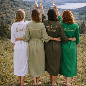 May include: Four people stand together outdoors, each wearing a different colored robe and holding a champagne glass. The robes are white, olive green, dark green, and light green. Gold text on the robes reads "Bride", "Bridesmaid", and "Mother of the bride".