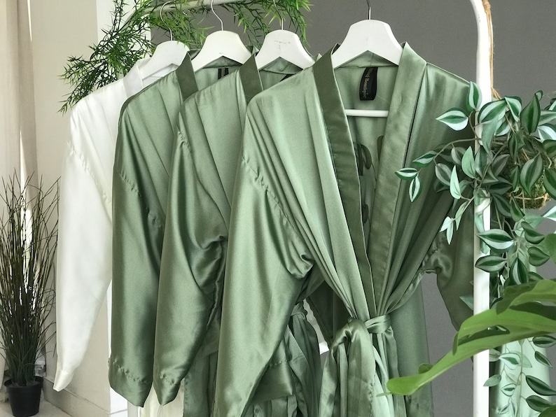 May include: Four satin robes hanging on white hangers. Three robes are sage green and one is white.
