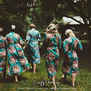 May include: Five women wearing teal floral print robes stand in a forest setting. The robes have a sash and are long-sleeved. The women are facing away from the camera.