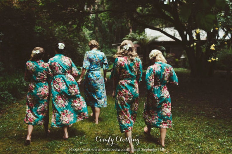 May include: Five women wearing teal floral print robes stand in a forest setting. The robes have a sash and are long-sleeved. The women are facing away from the camera.