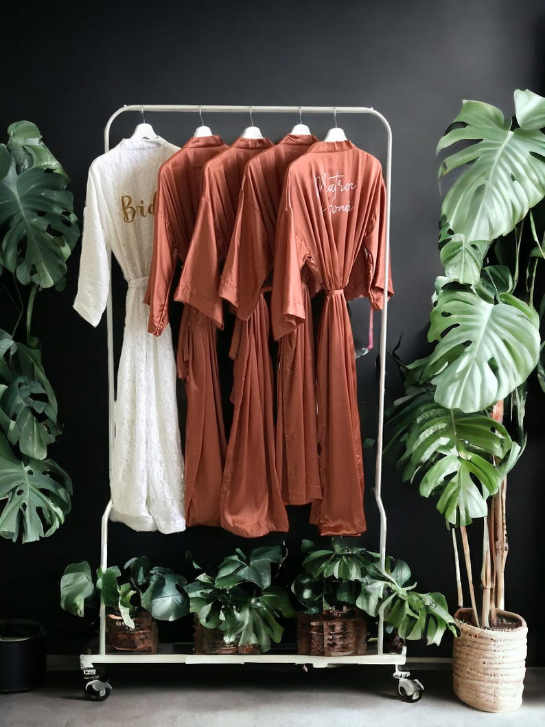 a collection of four robes hanging on a white rack, with two white robes and two red robes. The white robes have the word "Bride" printed on them, while the red robes have the word "Groom" printed on them. The rack is placed in front of a black wall, and there are several potted plants surrounding the rack, creating a cozy and inviting atmosphere.