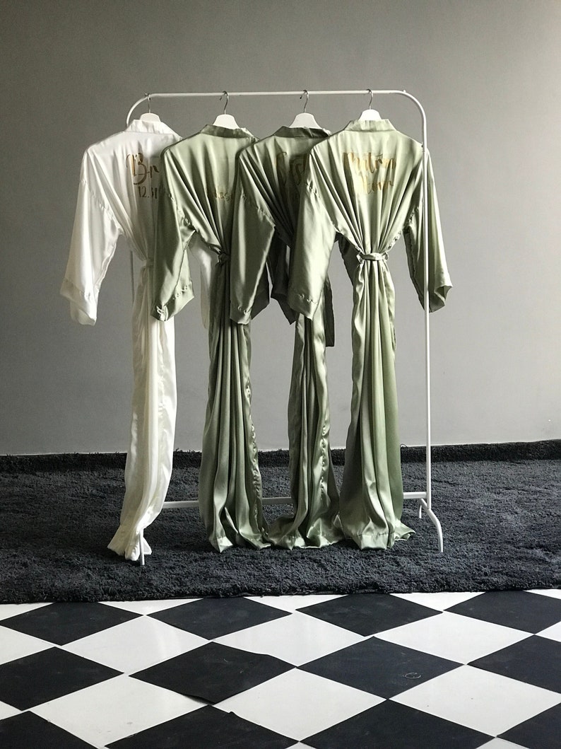 May include: Four robes hanging on a white rack. One robe is white with the text "Bride" in gold lettering. The other three robes are sage green with the text "Bride Tribe" in gold lettering.