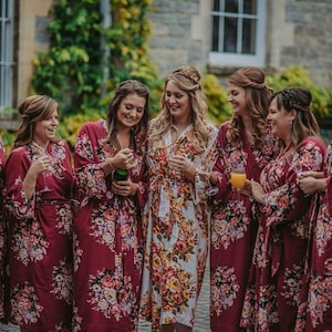May include: Five women wearing floral print robes in shades of burgundy and white. The women are smiling and holding glasses of champagne and orange juice.