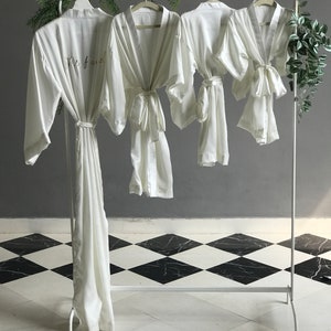 May include: Three white satin robes hanging on a white rack. The robe on the left has the text "Mrs. 2023" embroidered on the front. The robes are on a black and white checkered floor.