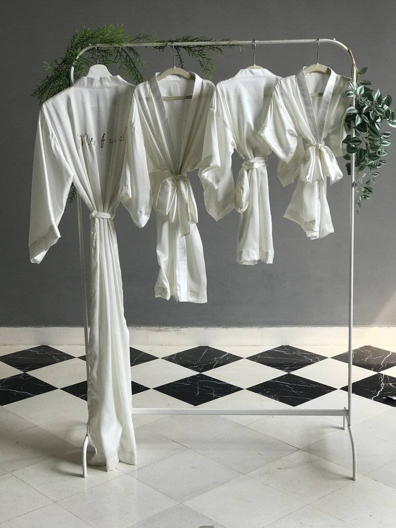 May include: Three white satin robes hanging on a white rack. The robe on the left has the text "Mrs. 2023" embroidered on the front. The robes are on a black and white checkered floor.