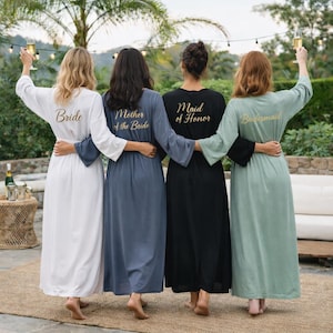 Pode incluir: Quatro roupões longos em branco, azul ardósia, preto e verde sálvia com letras douradas nas costas. O roupão branco diz Bride, o azul Mother of the Bride, o preto Maid of Honor e o verde Bridesmaid. Cada roupão possui mangas compridas.
