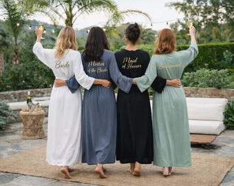 May include: Four long robes in white, slate blue, black, and sage green with gold script on the back. The white robe says Bride, the blue says Mother of the Bride, the black says Maid of Honor, and the green says Bridesmaid. Each robe has long sleeves.