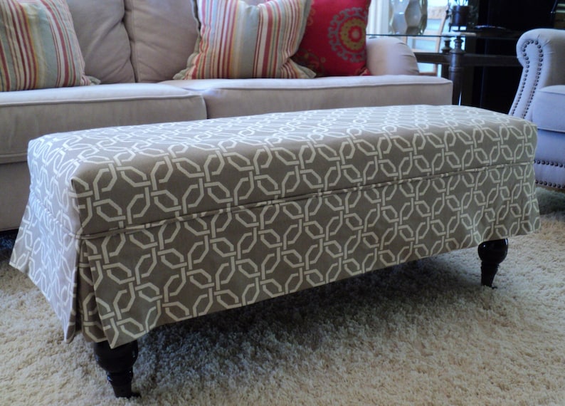 May include: A grey and white patterned ottoman with a skirt and four dark wood legs. The ottoman is sitting on a light beige rug.