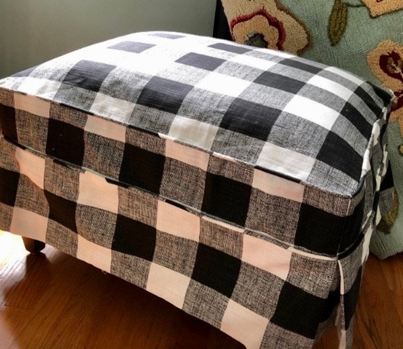 May include: Black and white checked fabric ottoman with a pleated skirt. The ottoman is made of a soft, textured fabric.