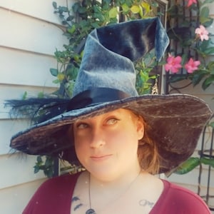 May include: A large, wide-brimmed witch hat with a tall, pointed crown. The hat is made of a dark gray fabric with a black ribbon around the base of the crown. The hat is decorated with a single black feather on the left side.