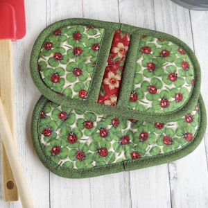 May include: A pair of green pot holders with a red and white floral pattern and a green border. The pot holders are made of fabric and have a clover and ladybug design.