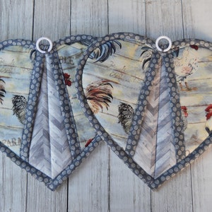 May include: Two heart-shaped pot holders with a rooster and farmhouse design. The pot holders are gray and white with a chevron pattern and a white ring on the top.