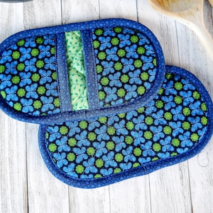 May include: Two blue and green floral patterned oval-shaped pot holders. The pot holders have a dark blue border and a light green patterned center. The background is a white wooden surface. Wooden spoons are in the background.