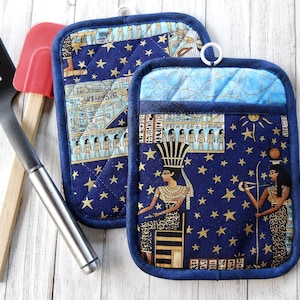 May include: Two blue quilted pot holders with a gold and blue Egyptian themed print. The print features stars, hieroglyphics, and figures of Egyptian royalty.